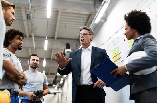 A manager in a suit gestures while talking to a team in an industrial space with whiteboards.