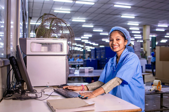 A smiling worker in a blue uniform and hairnet works at a computer in a brightly lit industrial facility.