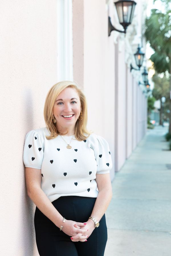 A person smiles while leaning against a light pink wall lined with outdoor lamps on a sidewalk.