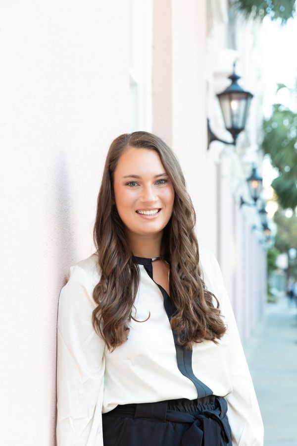 A smiling person with long, wavy brown hair wearing a white blouse with a black stripe, leaning against a pink wall.