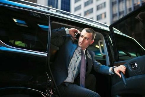 A person in a suit exits a black vehicle while talking on a mobile phone in a city setting.