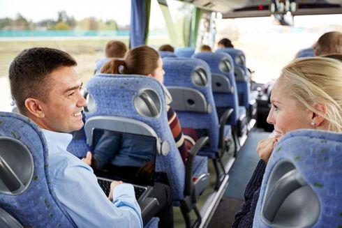 People sitting in blue seats on a bus, with two passengers in the foreground looking at each other and smiling.