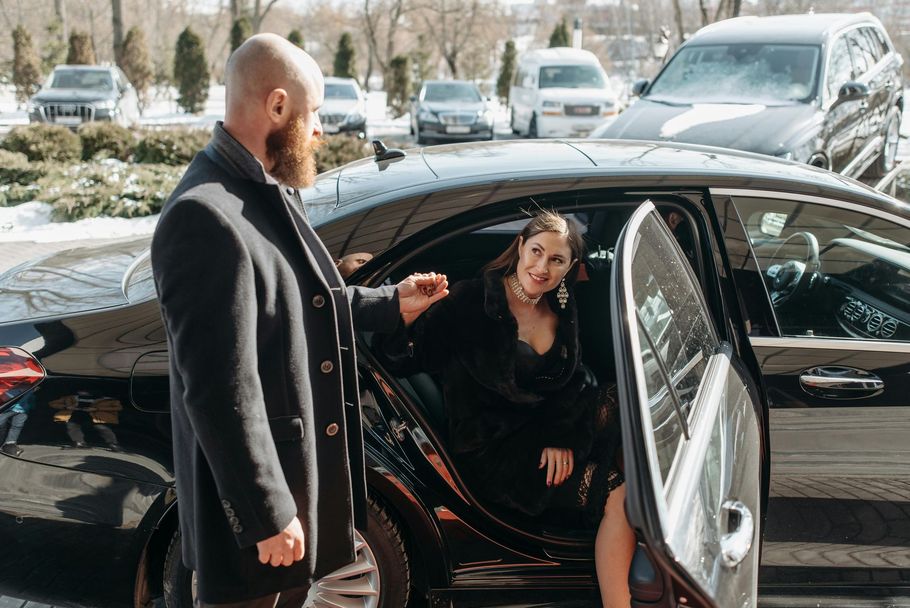 A bearded person helps a woman in a black dress exit a black sedan parked on a snowy lot.