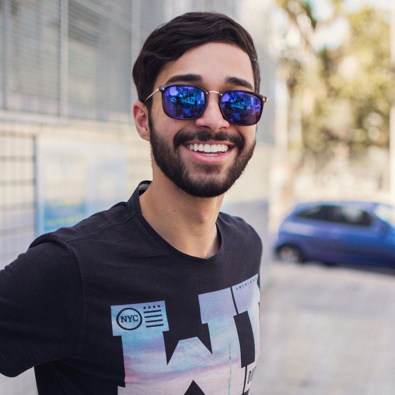 Man with sunglasses smiling outdoors, wearing black t-shirt. Blue car blurred in background.