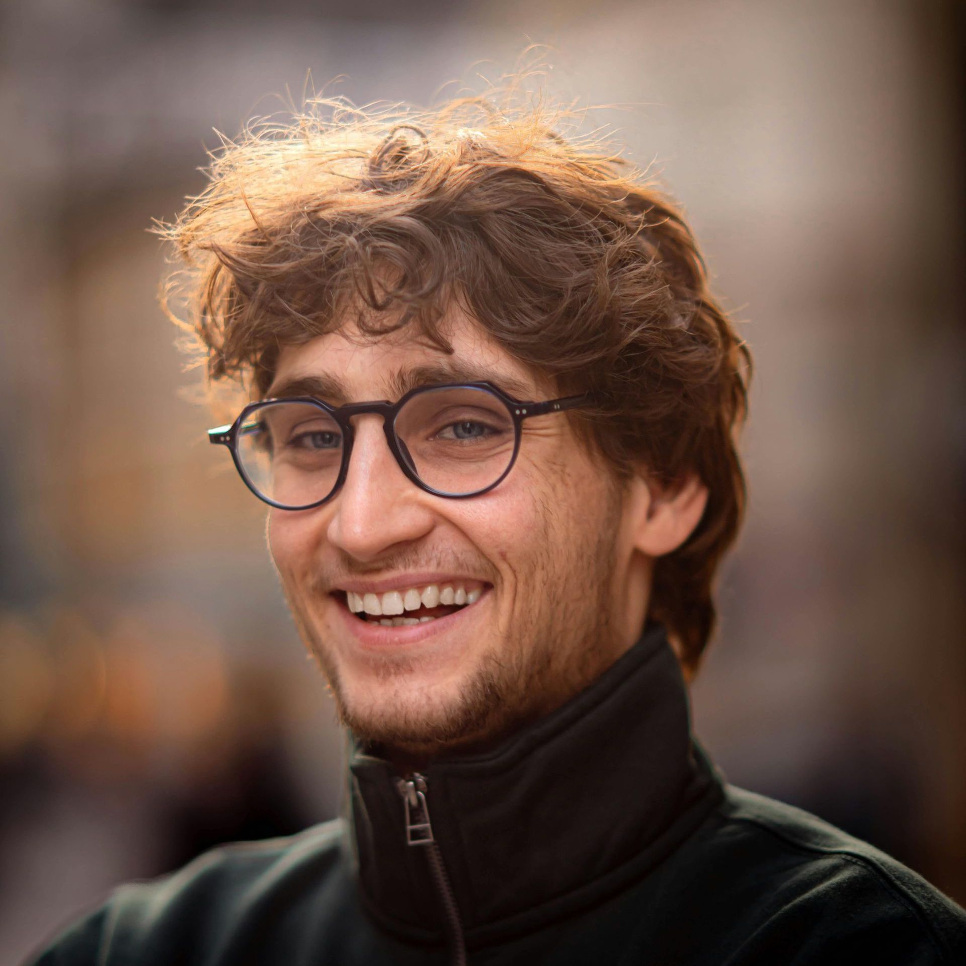 Man with curly brown hair and glasses smiles. Outdoors, wearing a dark jacket.