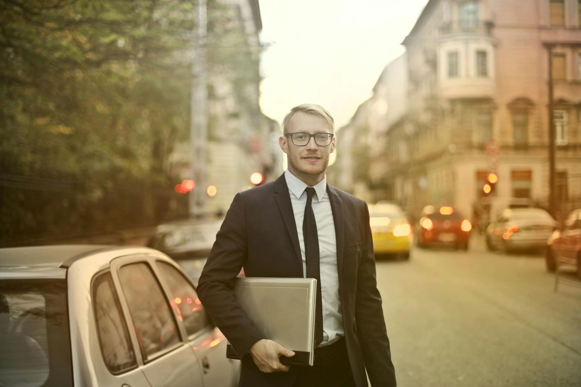 Man in suit holding a laptop, standing on a city street, smiling.