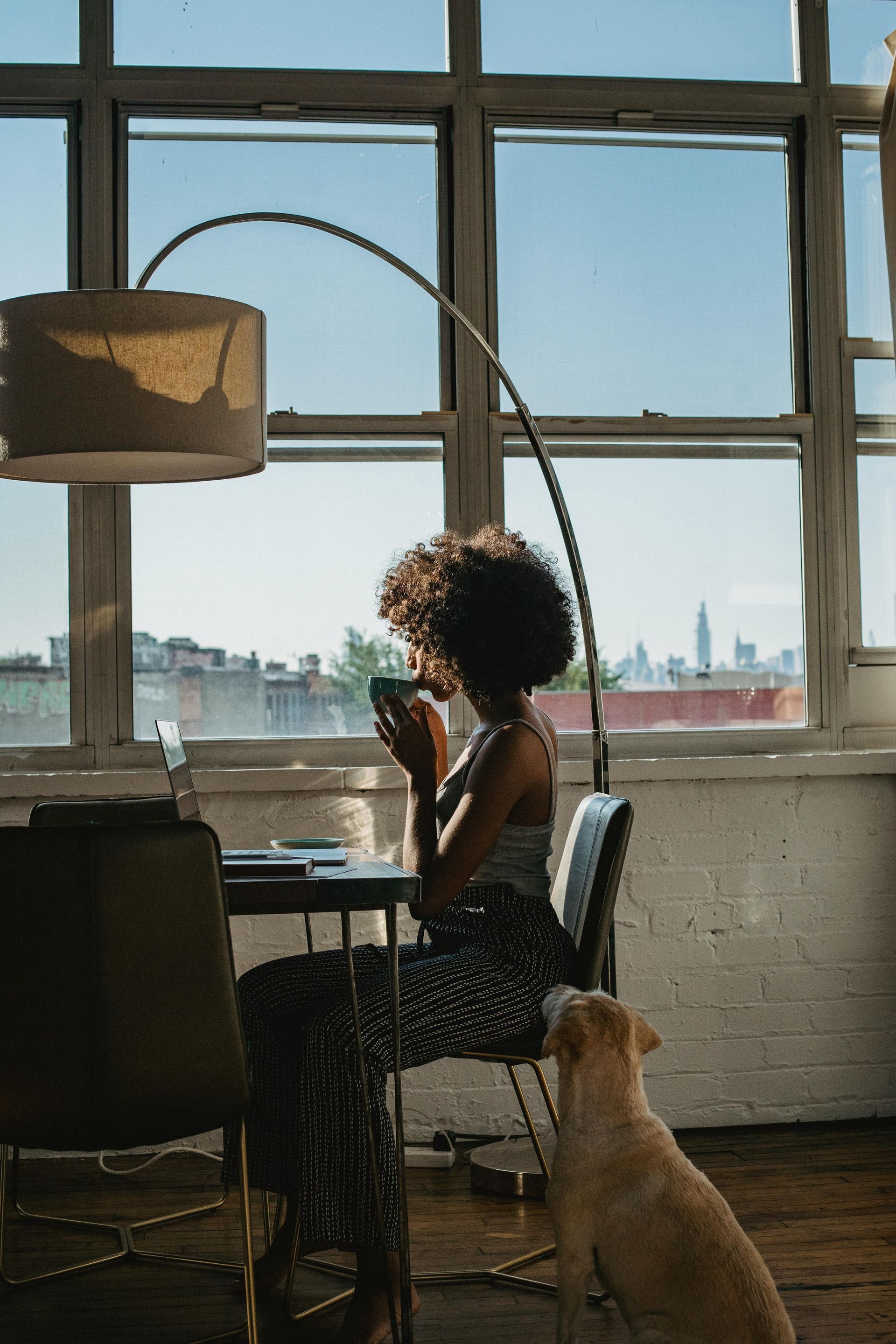 Woman sitting by window, drinking coffee, with a dog at her feet.