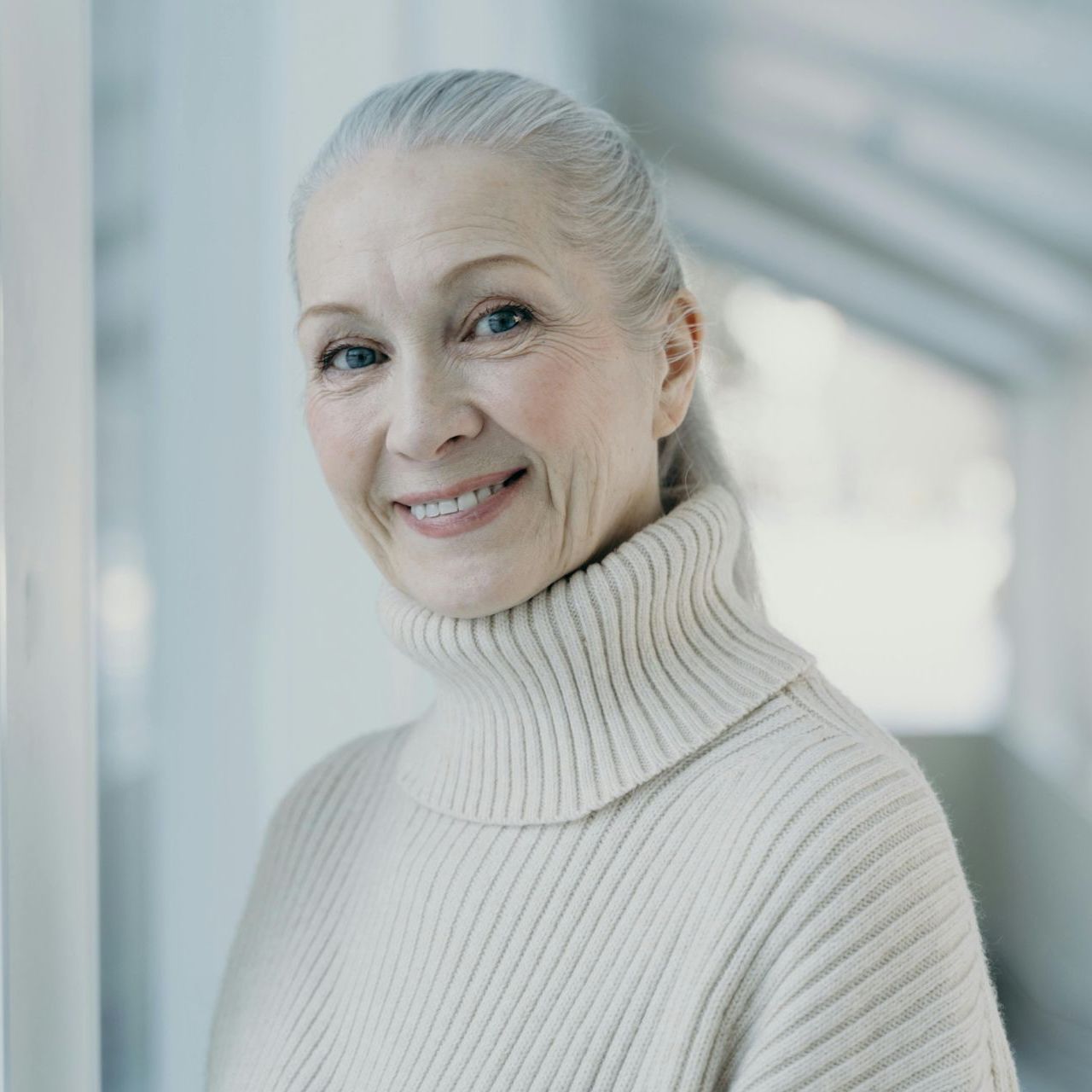 Woman with gray hair smiles, wearing a cream-colored turtleneck sweater, standing near a bright window.