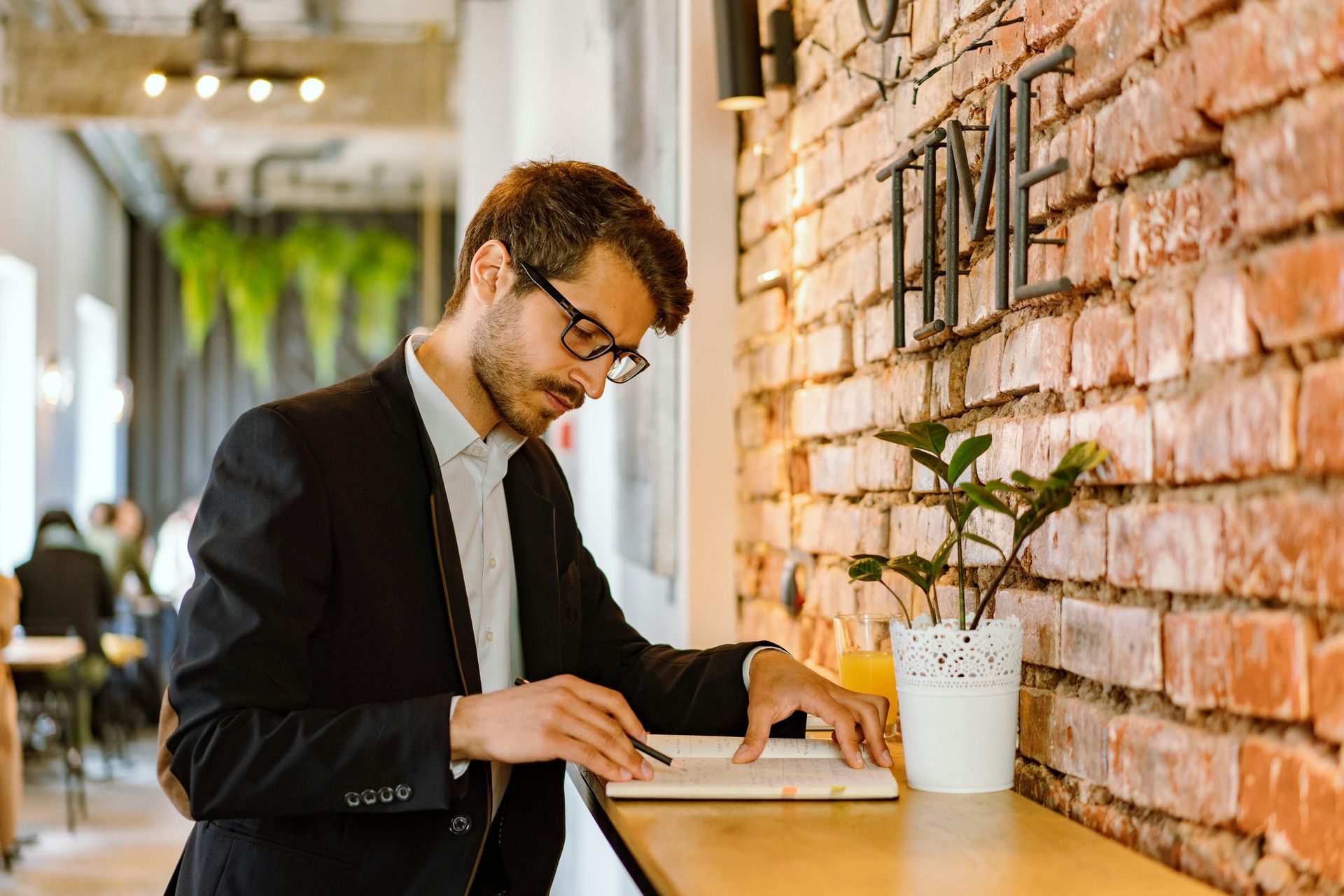 Man in glasses and suit jacket, writing in notebook at a brick wall counter in a cafe.
