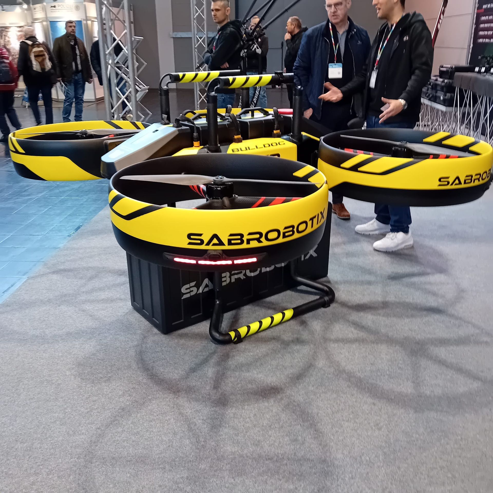 A yellow and black drone on display at a trade show, with several people observing.