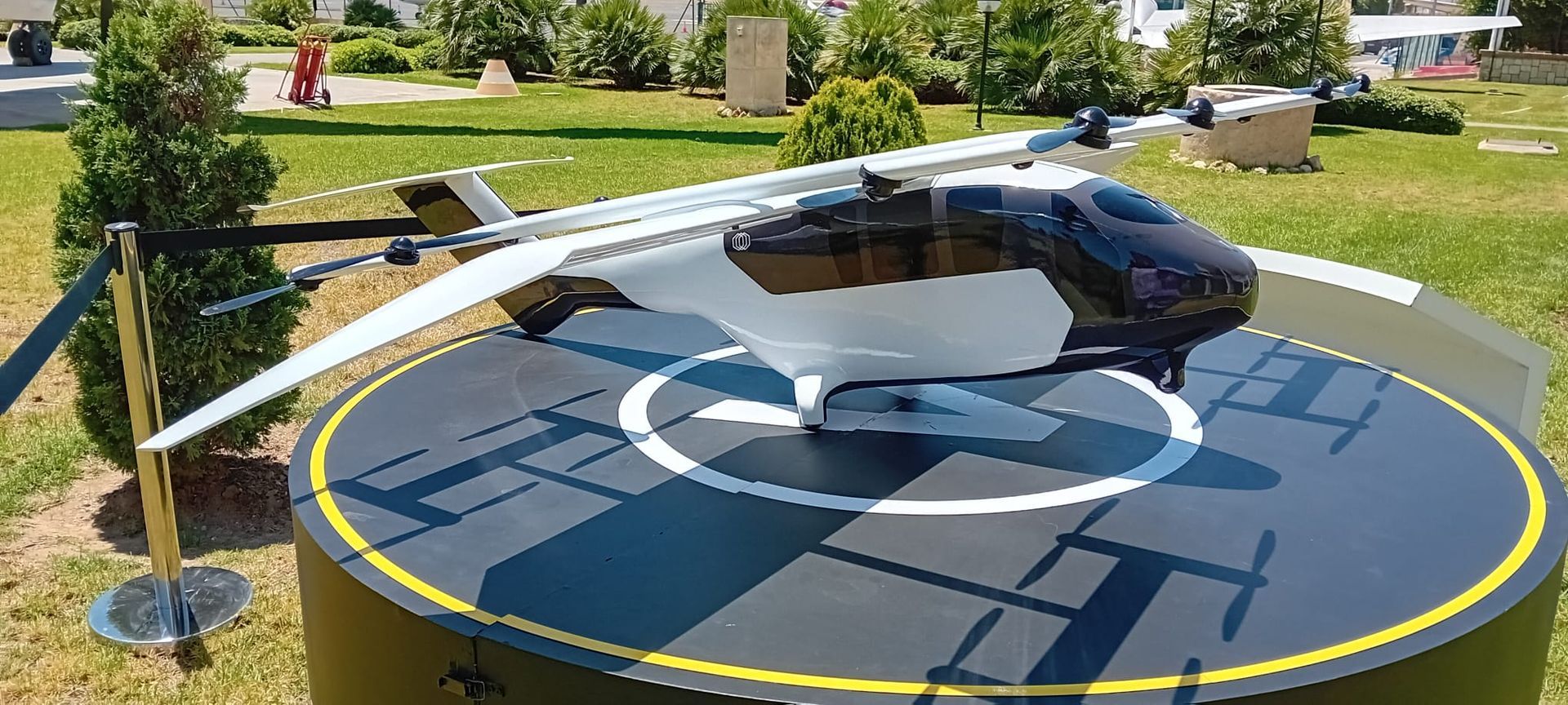A small white and black air taxi on a helipad outdoors with trees and grass.