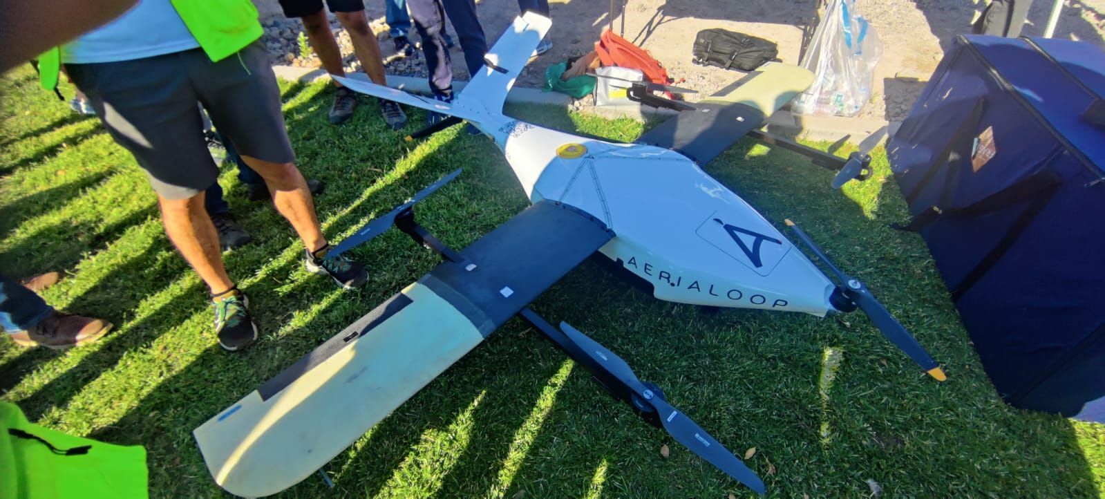 A white drone with black propellers and wings on green grass, with people surrounding it.