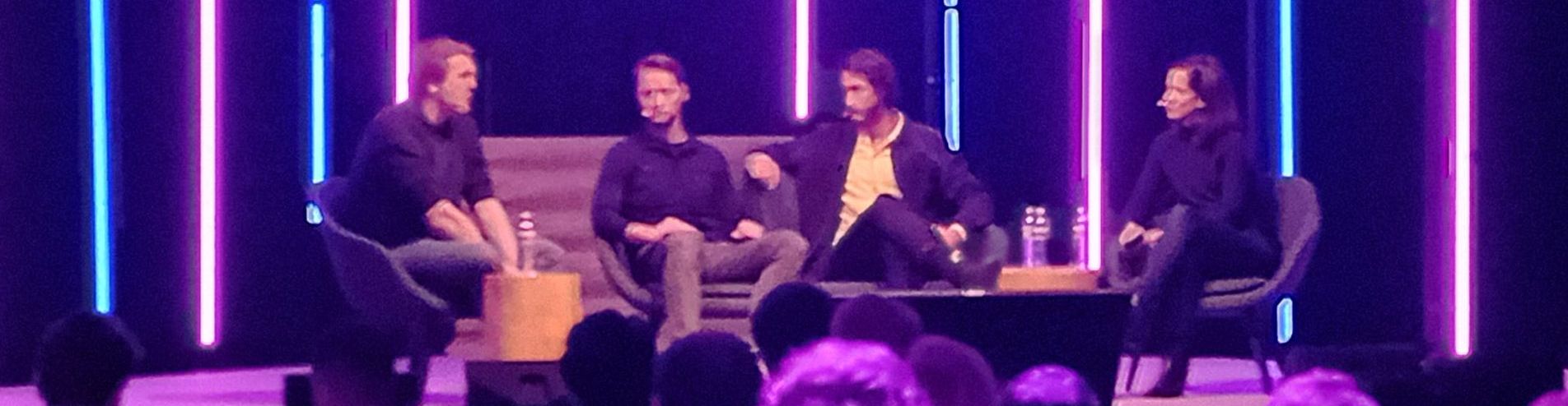Four people seated on a stage, possibly for a panel discussion. Lit by neon lights, with an audience in the foreground.