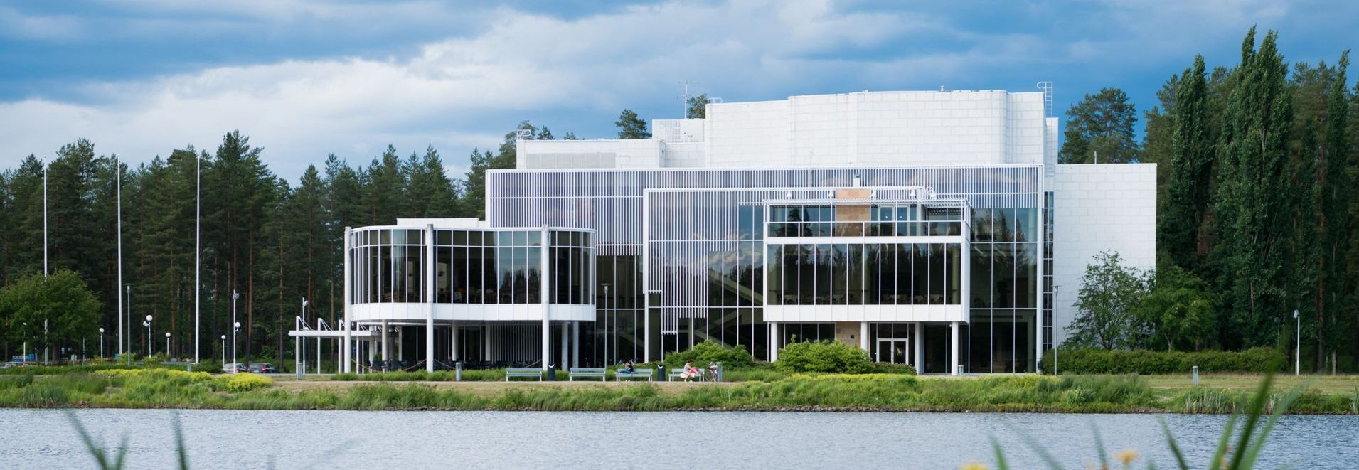 Modern white building with glass windows, by a lake and trees.