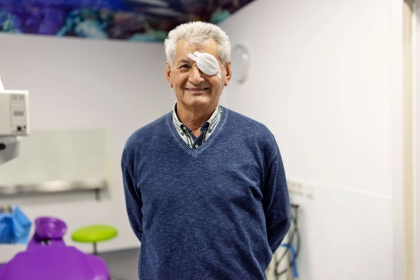 Un uomo con una benda sull'occhio sorride in uno studio medico. Indossa un maglione blu e una benda bianca sull'occhio.
