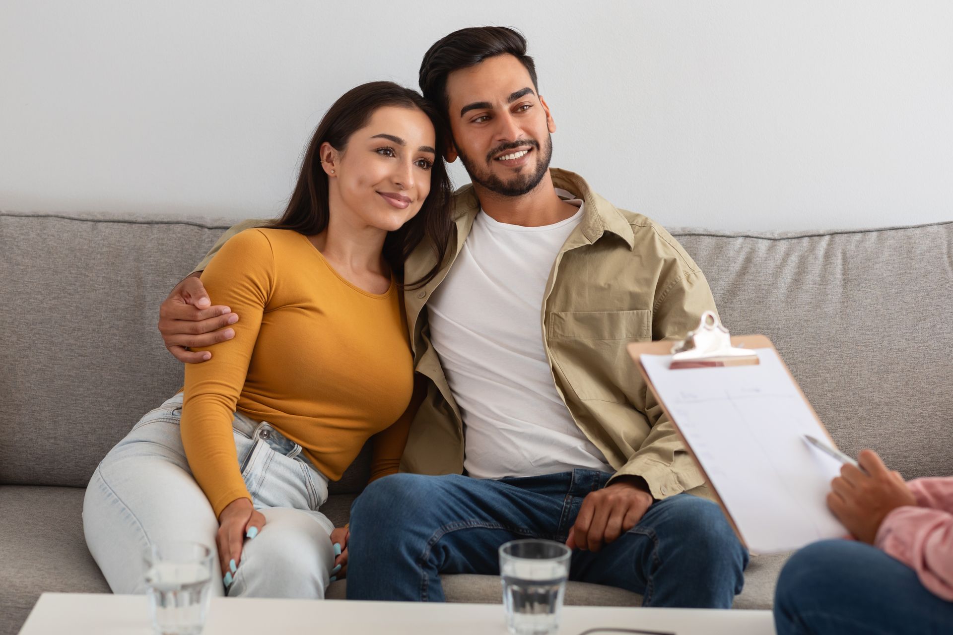 Couple sitting on a couch during a counseling session, smiling while a clipboard is held in front of them