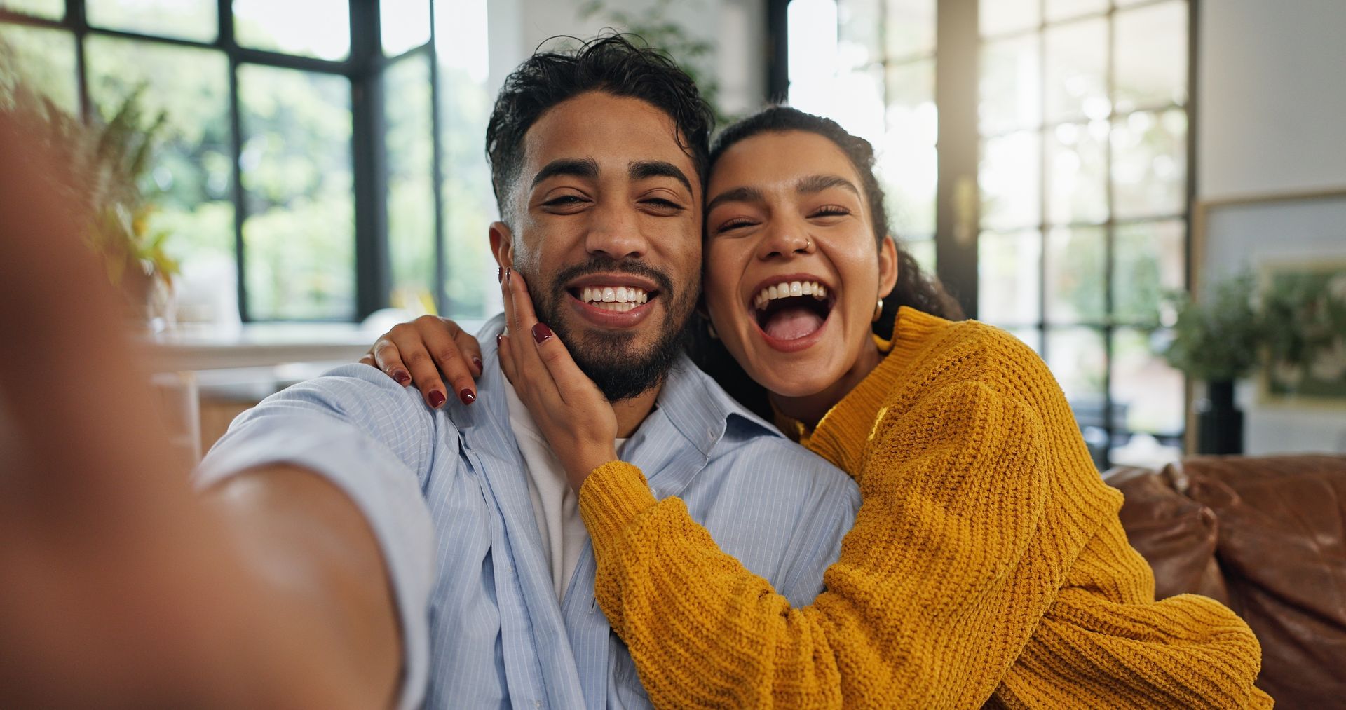 Two smiling people taking a selfie indoors, one in a mustard sweater, with large windows behind them.