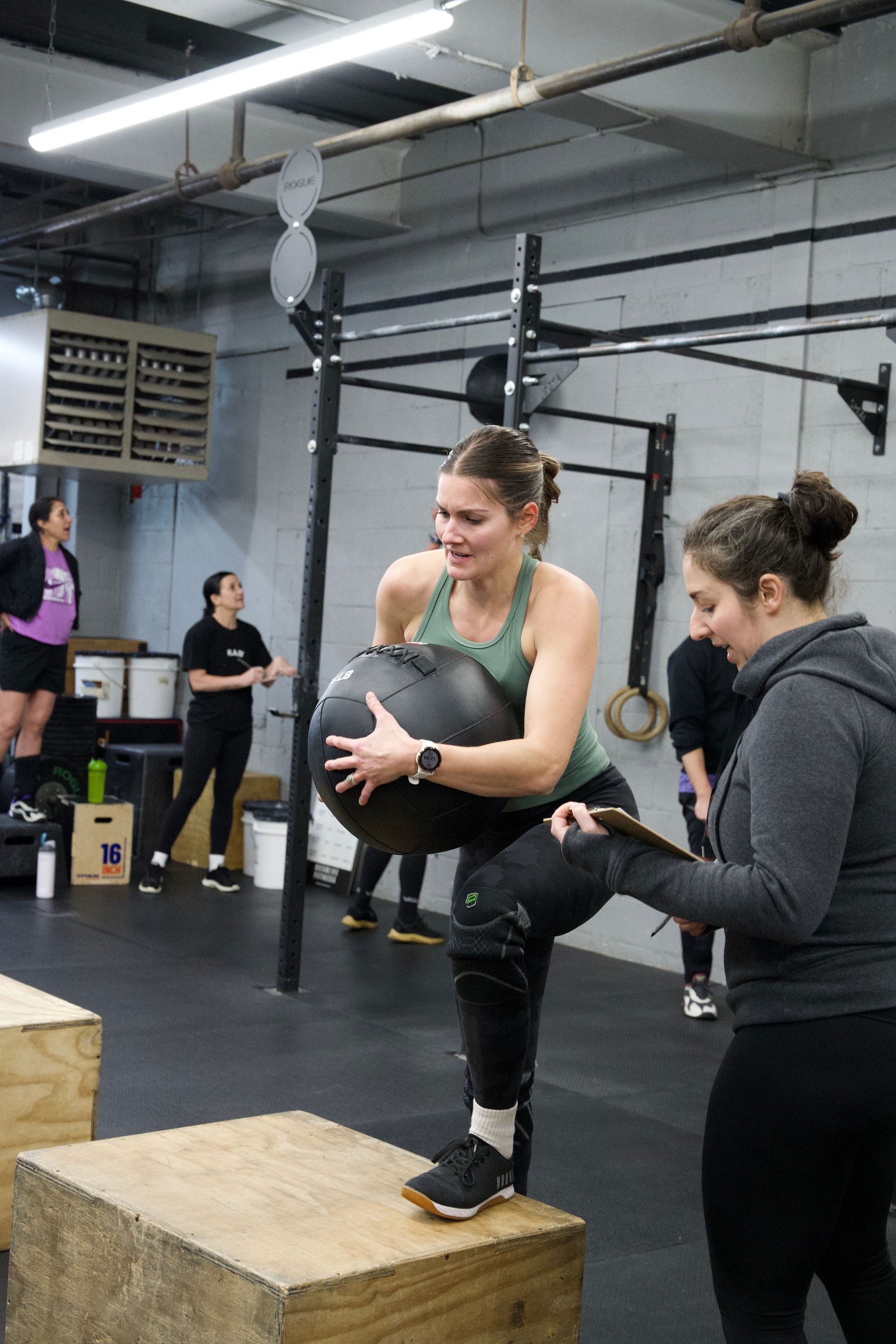 Woman balancing on a box holding a medicine ball in a gym, with a trainer beside her.