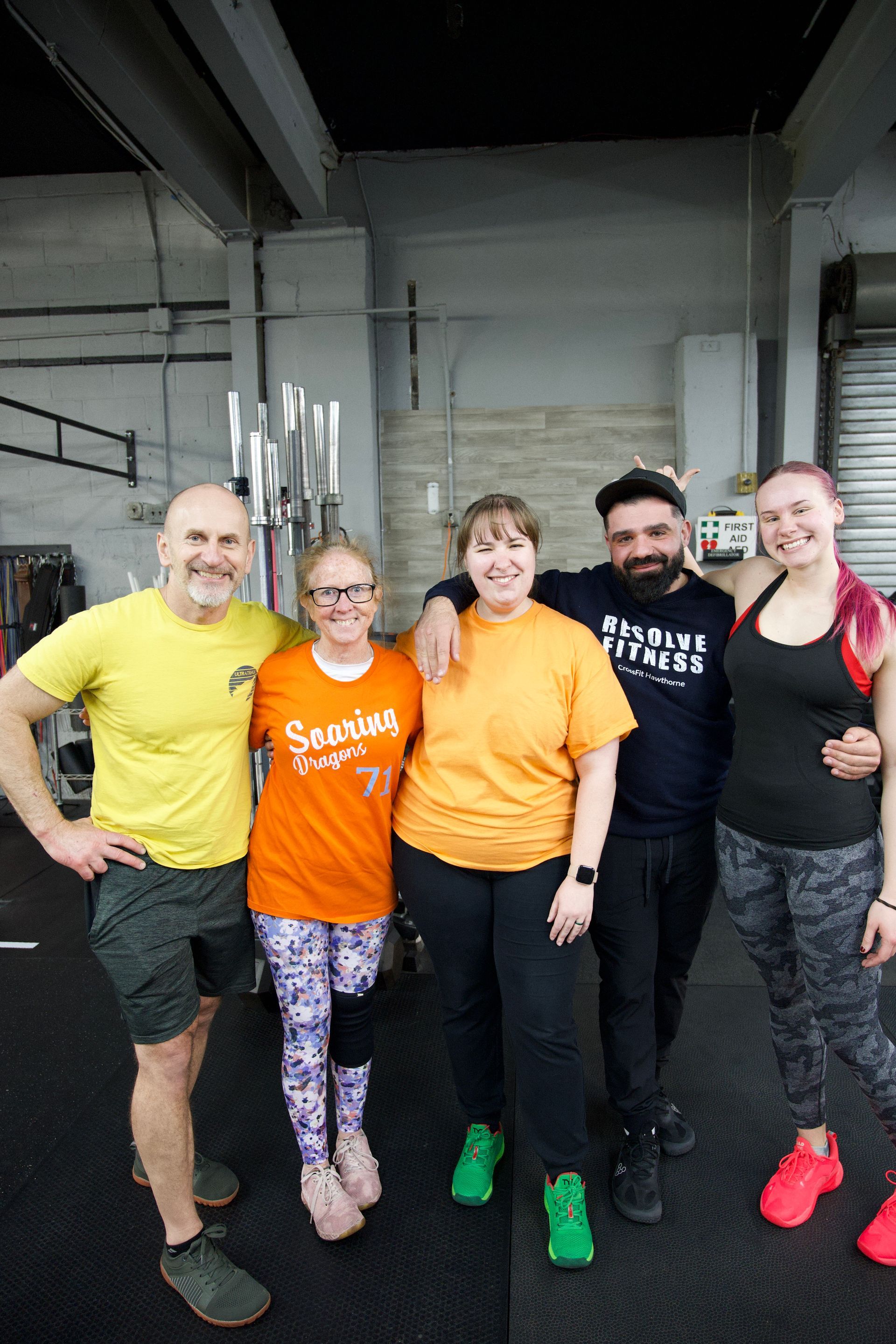 Five people posing together in a gym, smiling in colorful workout clothes.