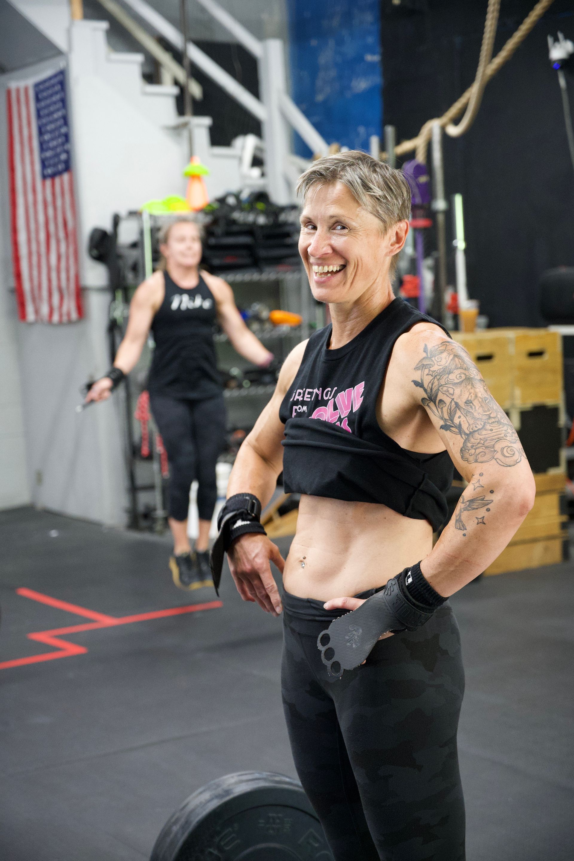 Smiling athlete in black workout gear stands in a gym with ropes and a U.S. flag in the background
