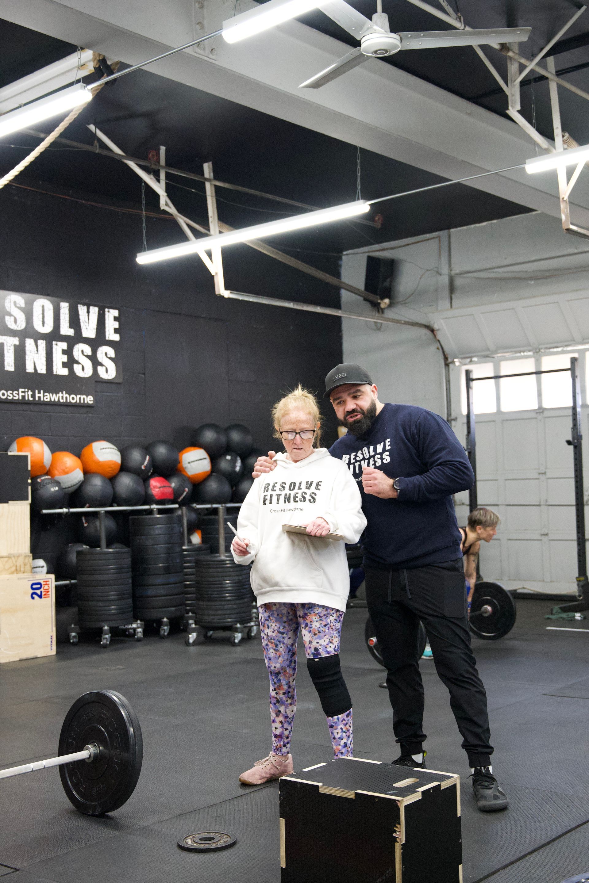 Two people in a gym stand beside a barbell and a plyo box, reviewing a phone together.