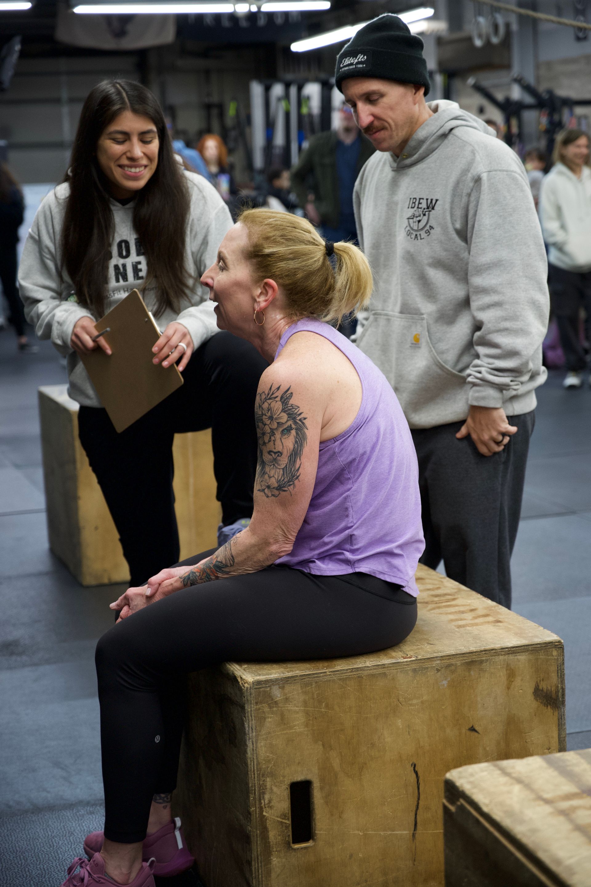 Woman with purple top sitting on a wooden box, talking with two people in a gym or warehouse space