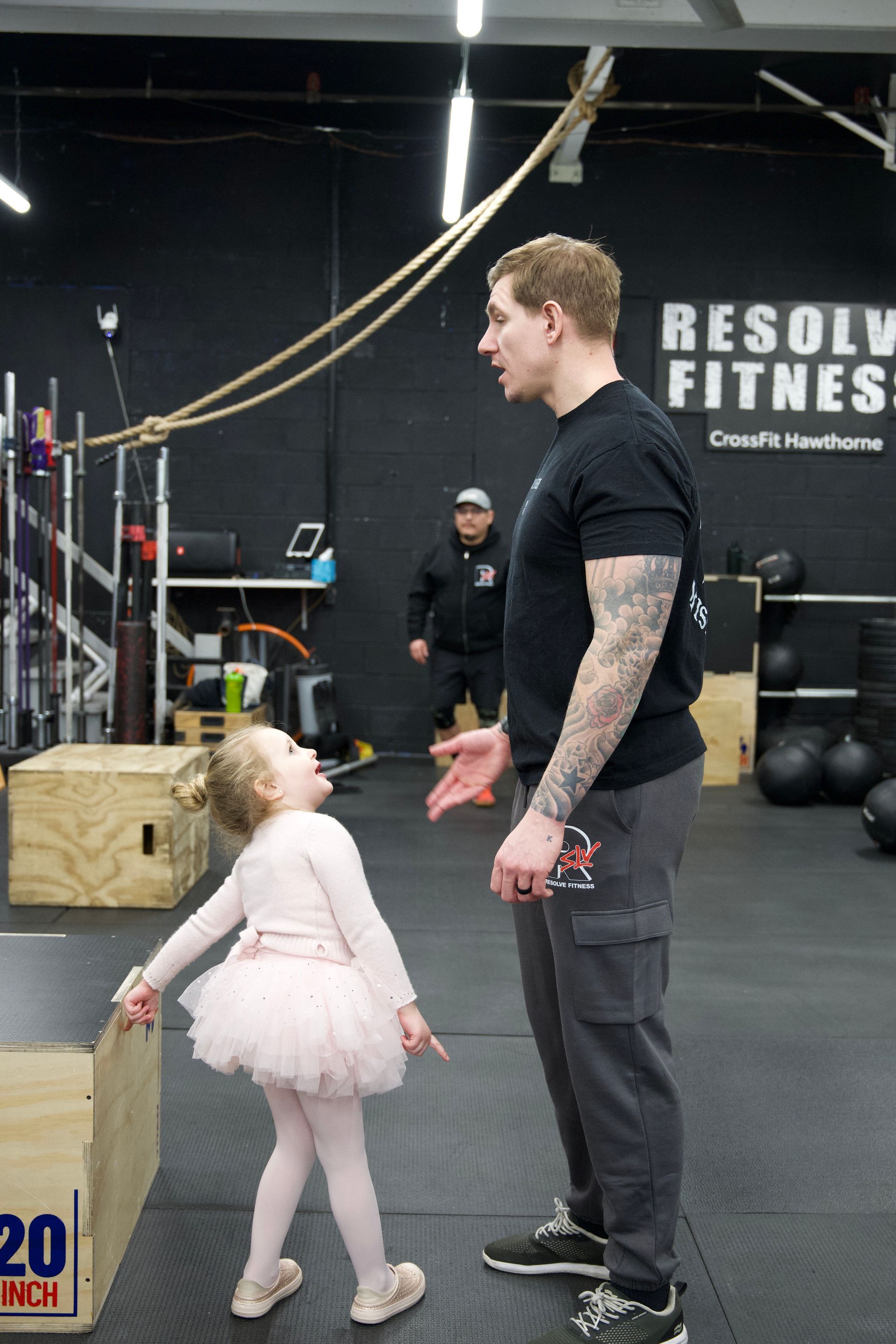Man and small child standing in a gym, facing each other near workout equipment and a wooden box