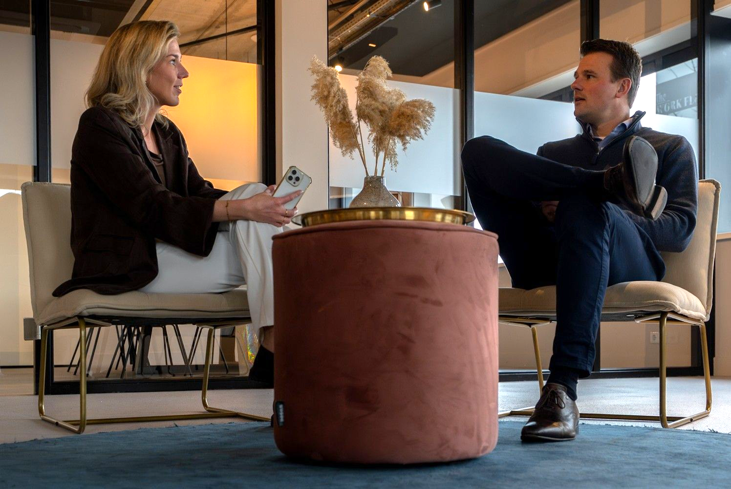 Twee mensen zitten in een moderne lounge en praten met elkaar aan een kleine ronde tafel met gedroogde bloemen.