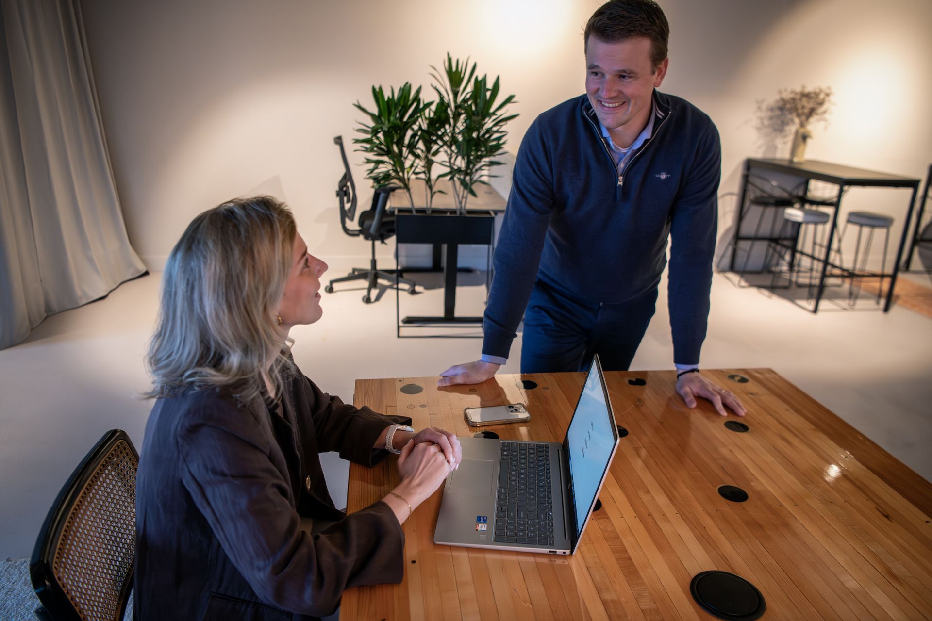 Twee mensen in gesprek aan een houten tafel met een laptop in een lichte kantoorruimte.