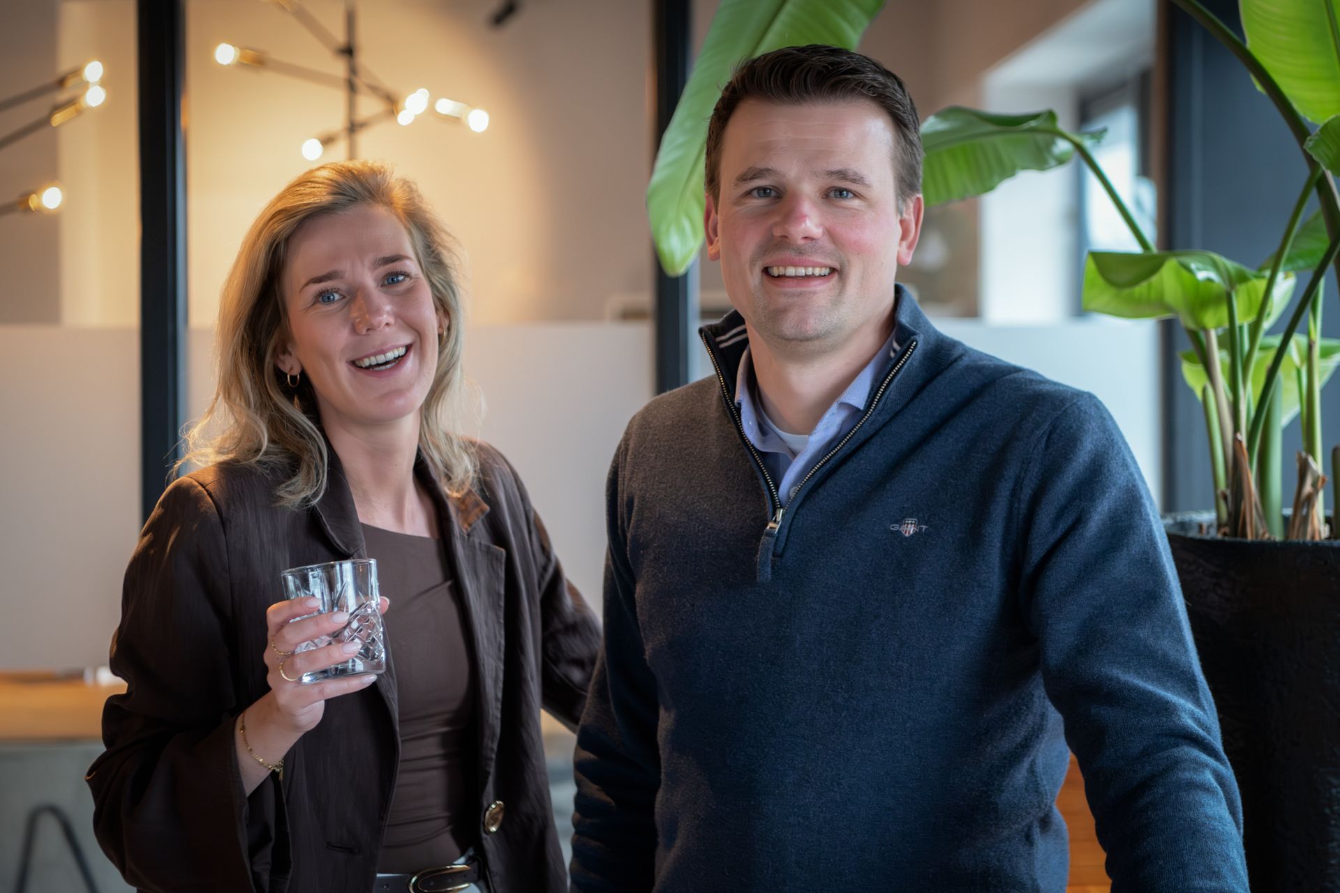 Twee lachende collega's staan ​​binnen, een van hen met een glas in de hand, in een sfeervol verlichte kantoorlounge.