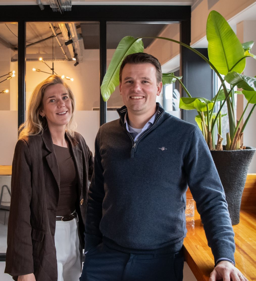 Twee mensen staan in een moderne kantoorlobby naast een grote potplant en glimlachen naar de camera.