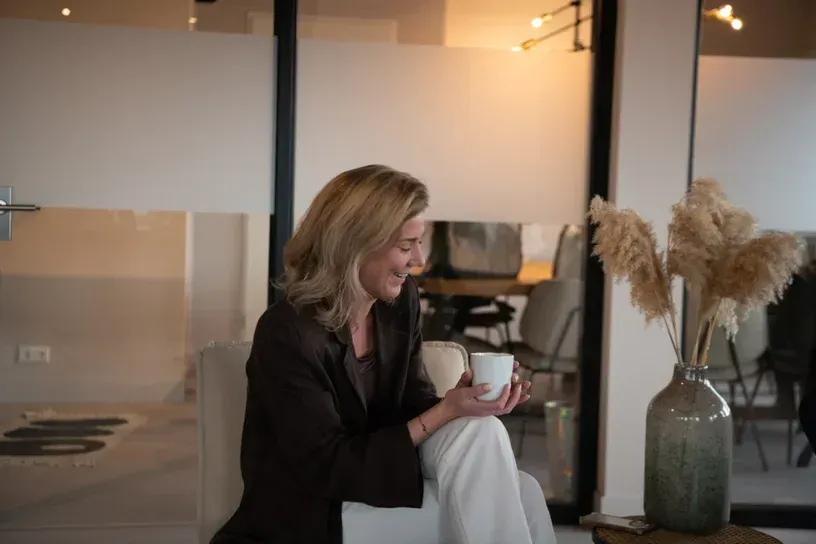 Een vrouw zit lachend op een bank, met een mok in haar hand, in een gezellige, moderne kamer.