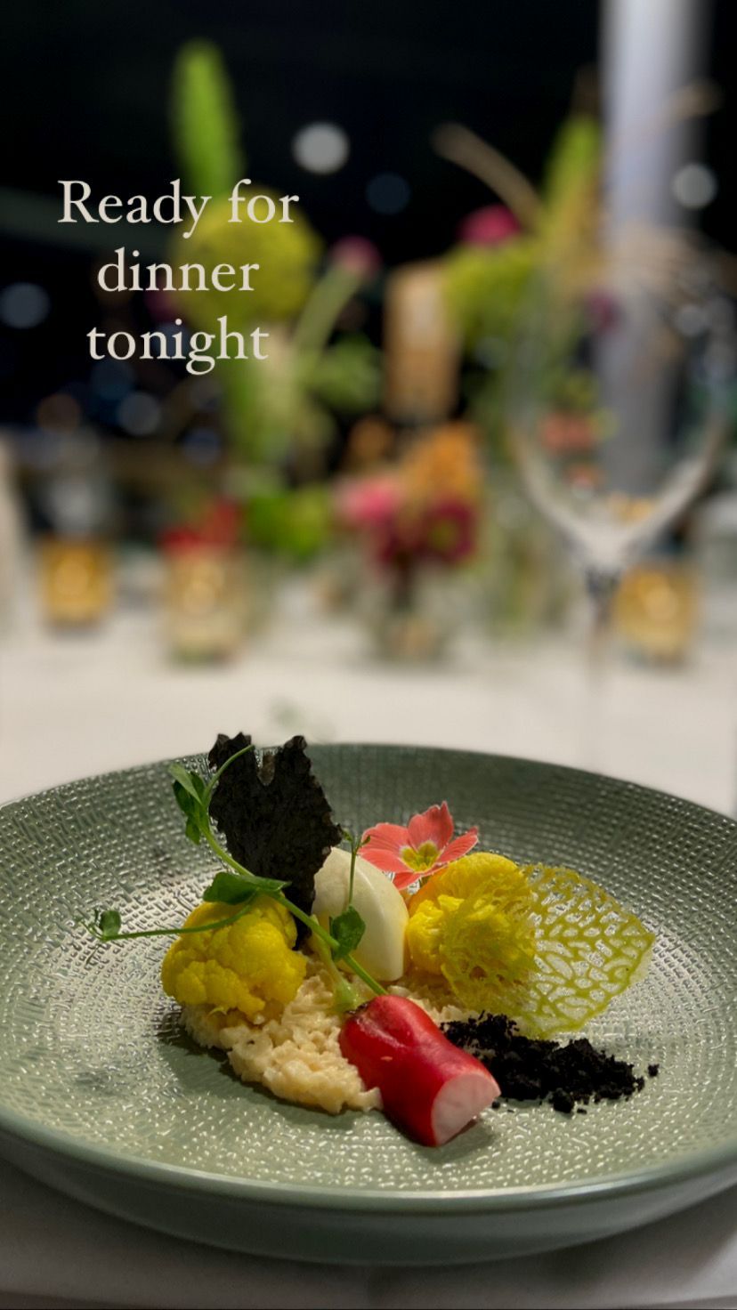 Een verfijnd vega gerecht gepresenteerd op een groen bord met een subtiele textuur. Het gerecht bevat gele bloemkoolroosjes, een rode radijs, zwarte crumble, een luchtige witte mousse en een decoratieve groene krokante tuile. Verder is het gerecht afgewerkt met een takje verse erwtenscheuten en een roze eetbare bloem, wat een kleurrijk en harmonieus geheel creëert.