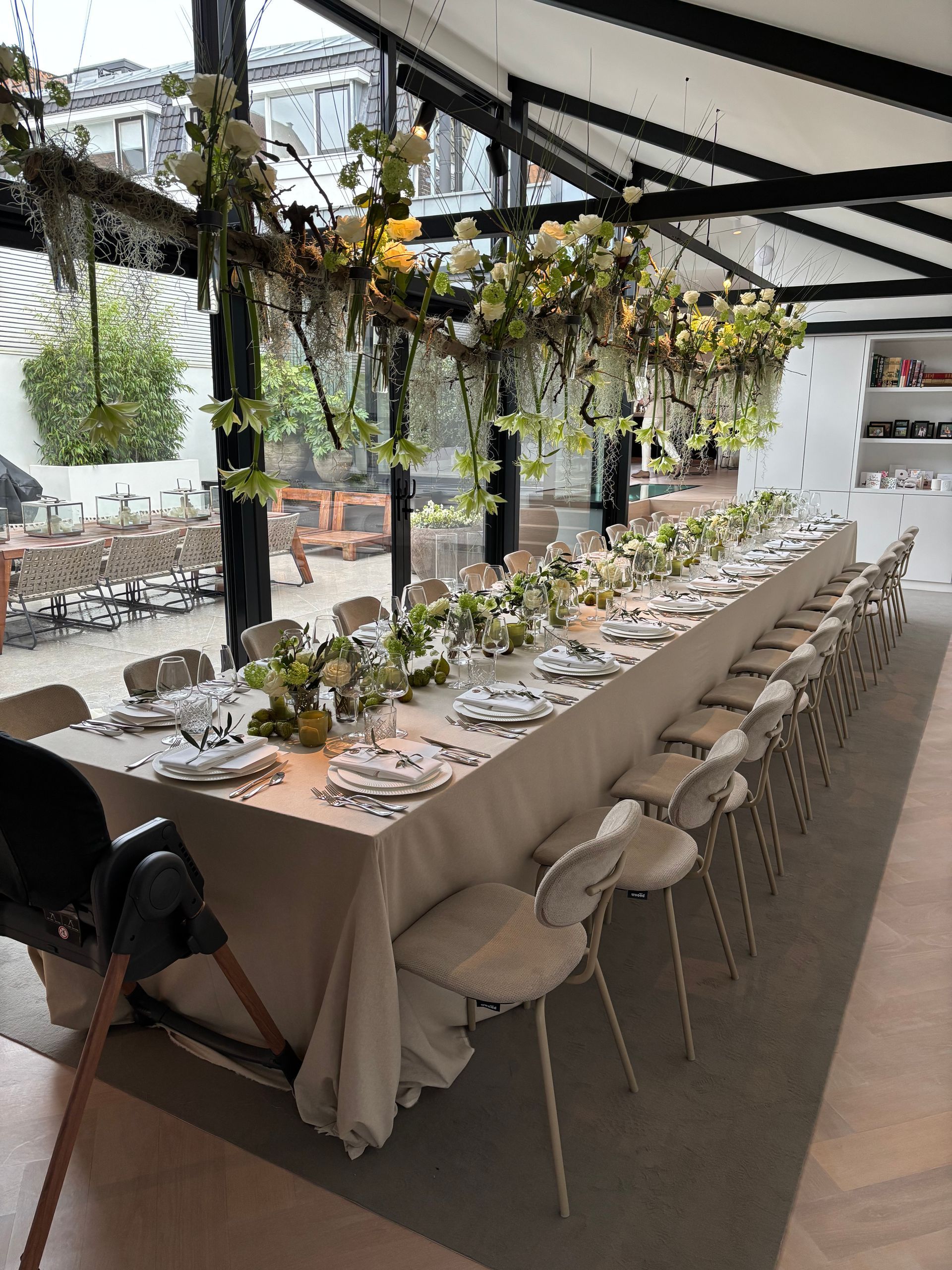 Lange gedekte tafel met hangende bloemendecoratie en witte stoelen in een lichte eventlocatie