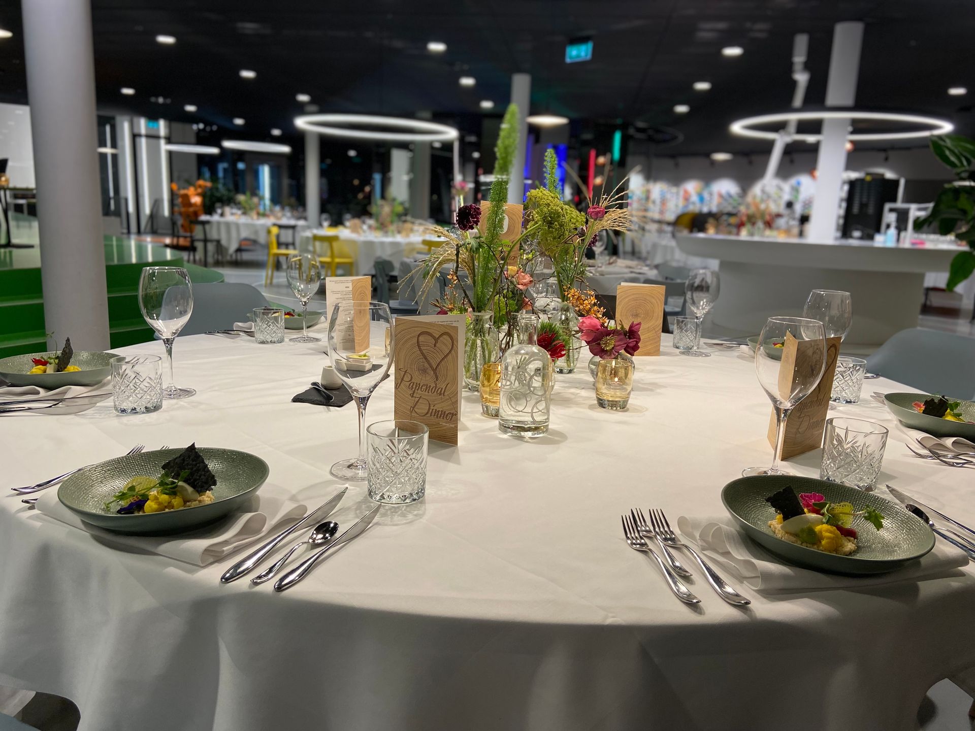 Een elegant gedekte dinertafel met een witte tafelkleed, omringd door glazen en porseleinen borden. Op de tafel staan diverse decoratieve elementen, zoals glazen vazen met kleurrijke wilde bloemen en houten menukaarten met een hartmotief en de tekst