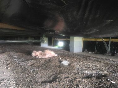 Inside a dark crawlspace, a dirt mound with pink insulation sits beneath a low floor beam.