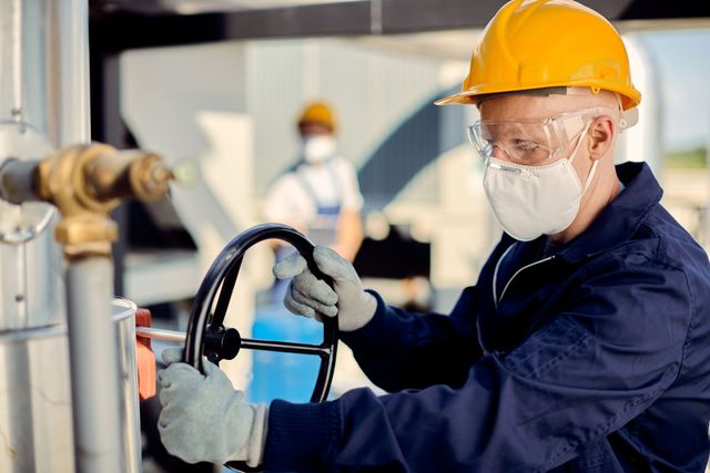 Worker in yellow hard hat and mask operating industrial equipment in a factory.