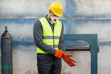 Worker in yellow hard hat, neon vest, and mask adjusting orange gloves beside industrial machinery.