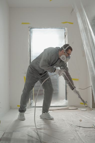 Person in a respirator using a long tool to clean or scrape a room under renovation.