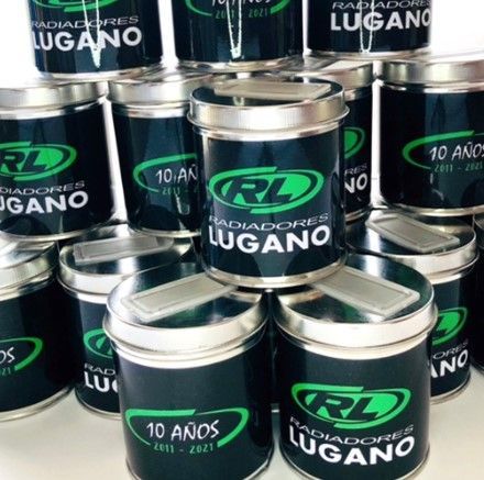 Latas apiladas de metal negro con las etiquetas “Lugano” y “10 años”, con tapas plateadas y detalles verdes.