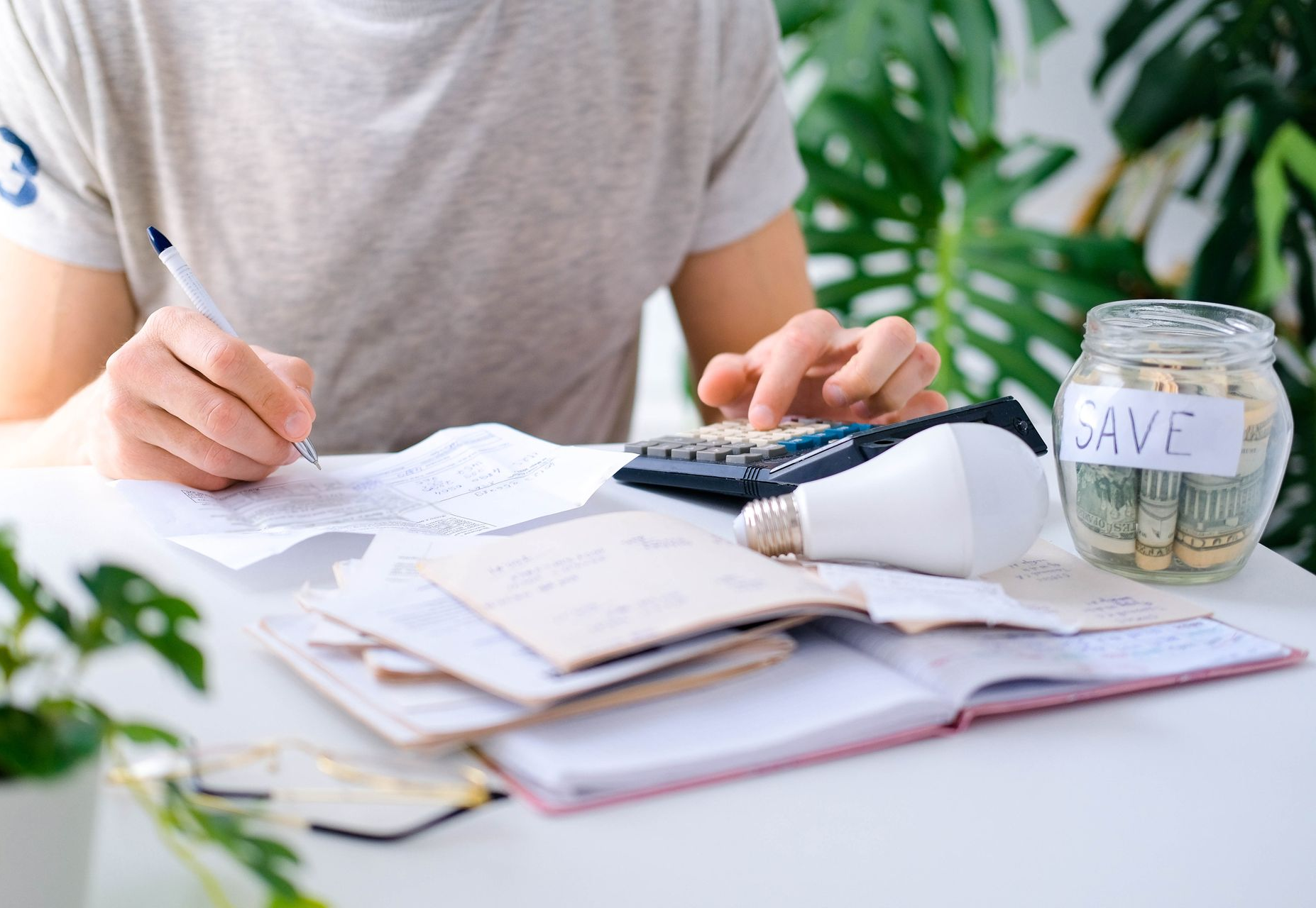 Person calculating finances with a calculator, receipts, and a savings jar on a table.