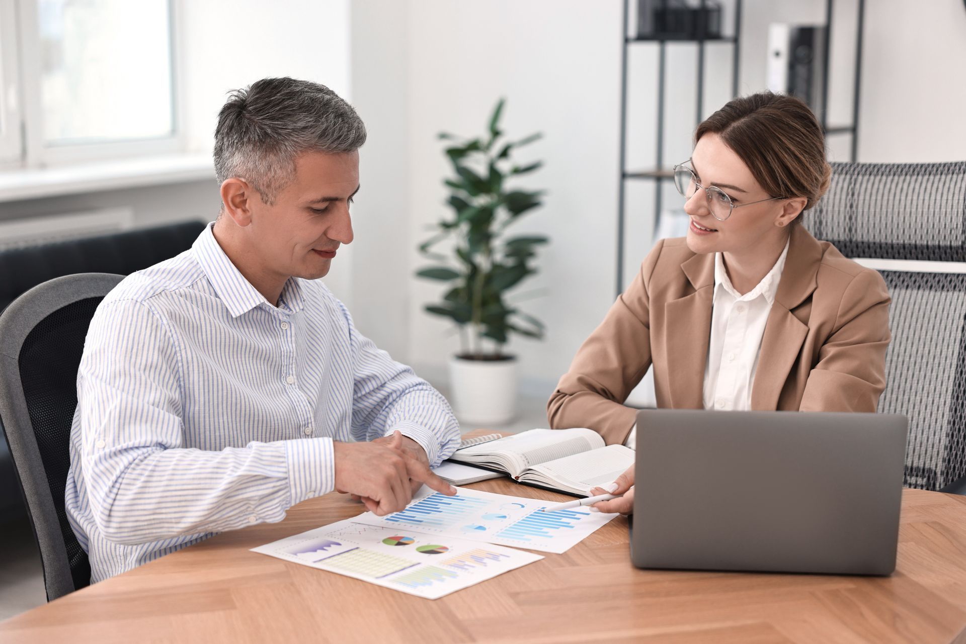 Man and woman reviewing documents at a table with laptop, plant, and notepad in an office setting.