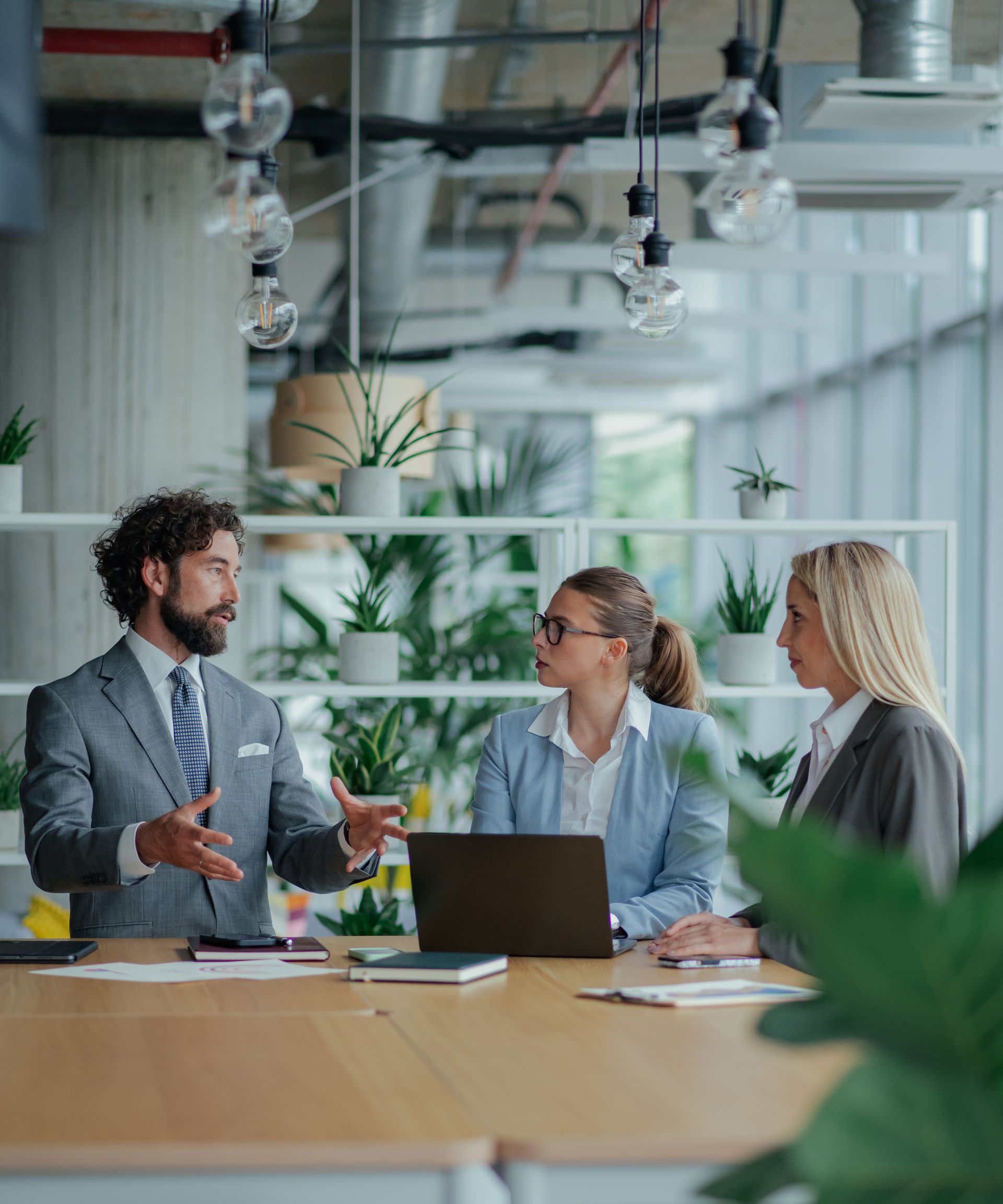 Three business people in suits at a table. Man with beard gestures, two women listen, laptop open, plants in background.