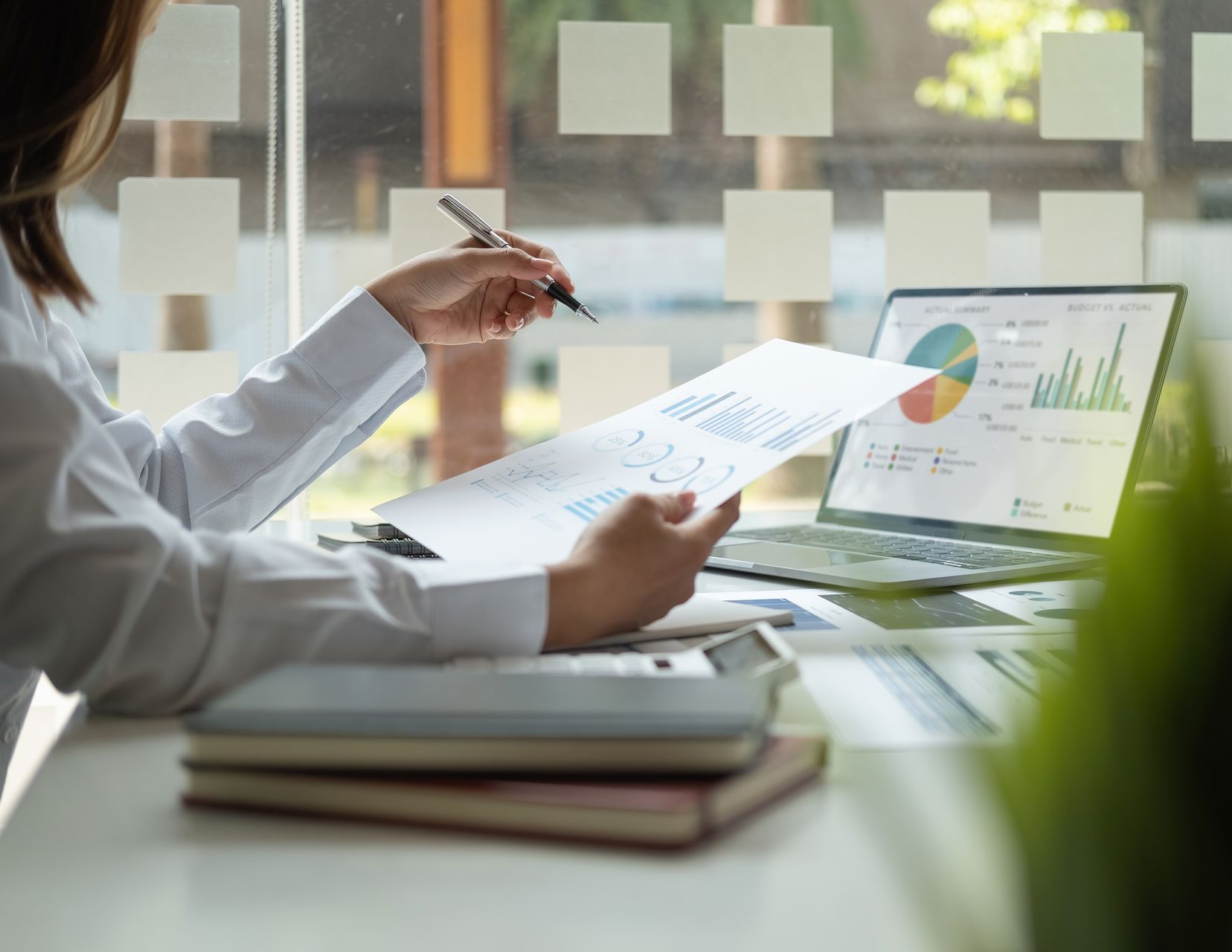 Woman reviews financial documents, pointing with a pen, laptop with charts in the background.