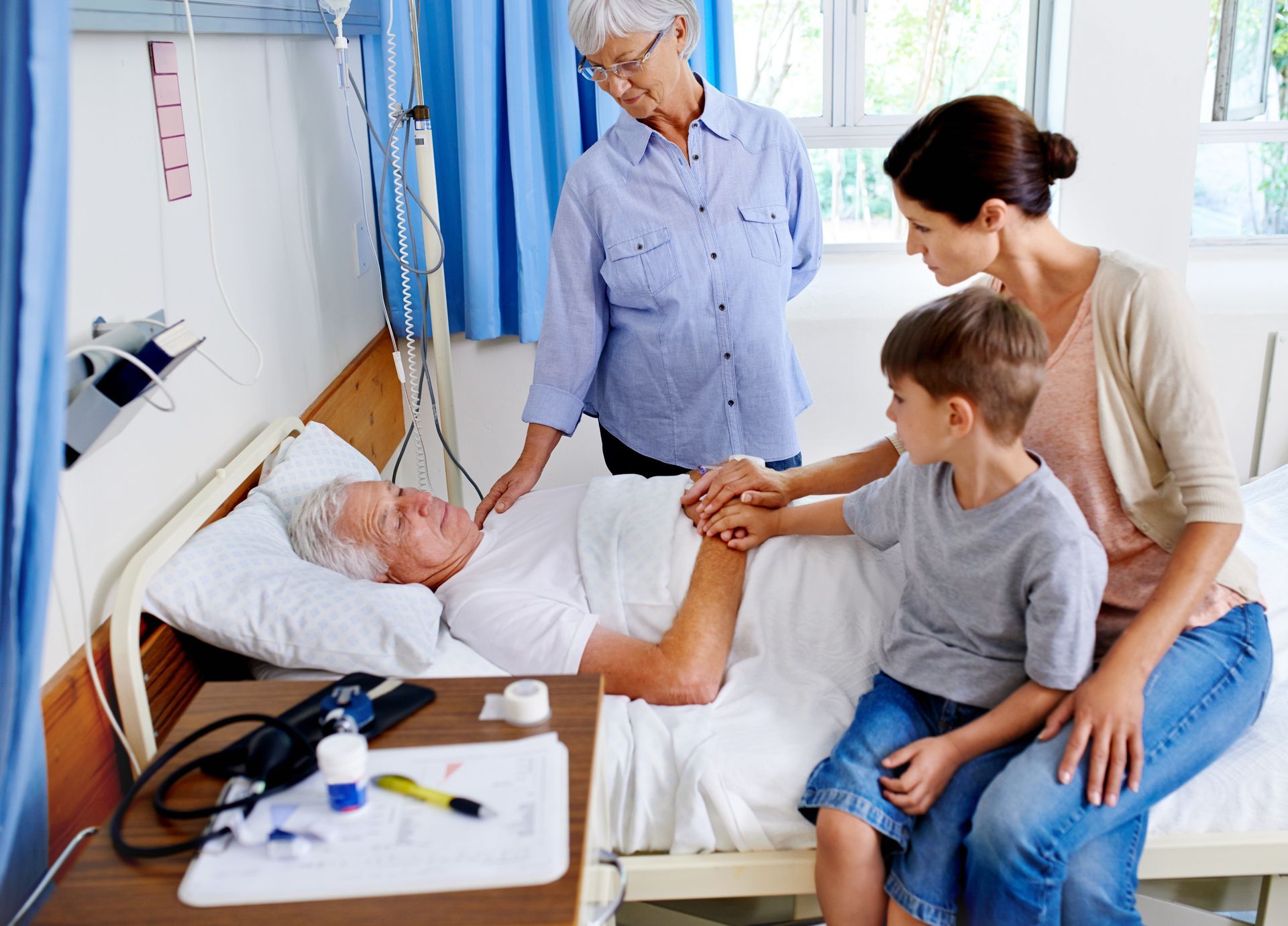 Family visiting a person in a hospital bed; grandmother, mother, and child touch his hand.