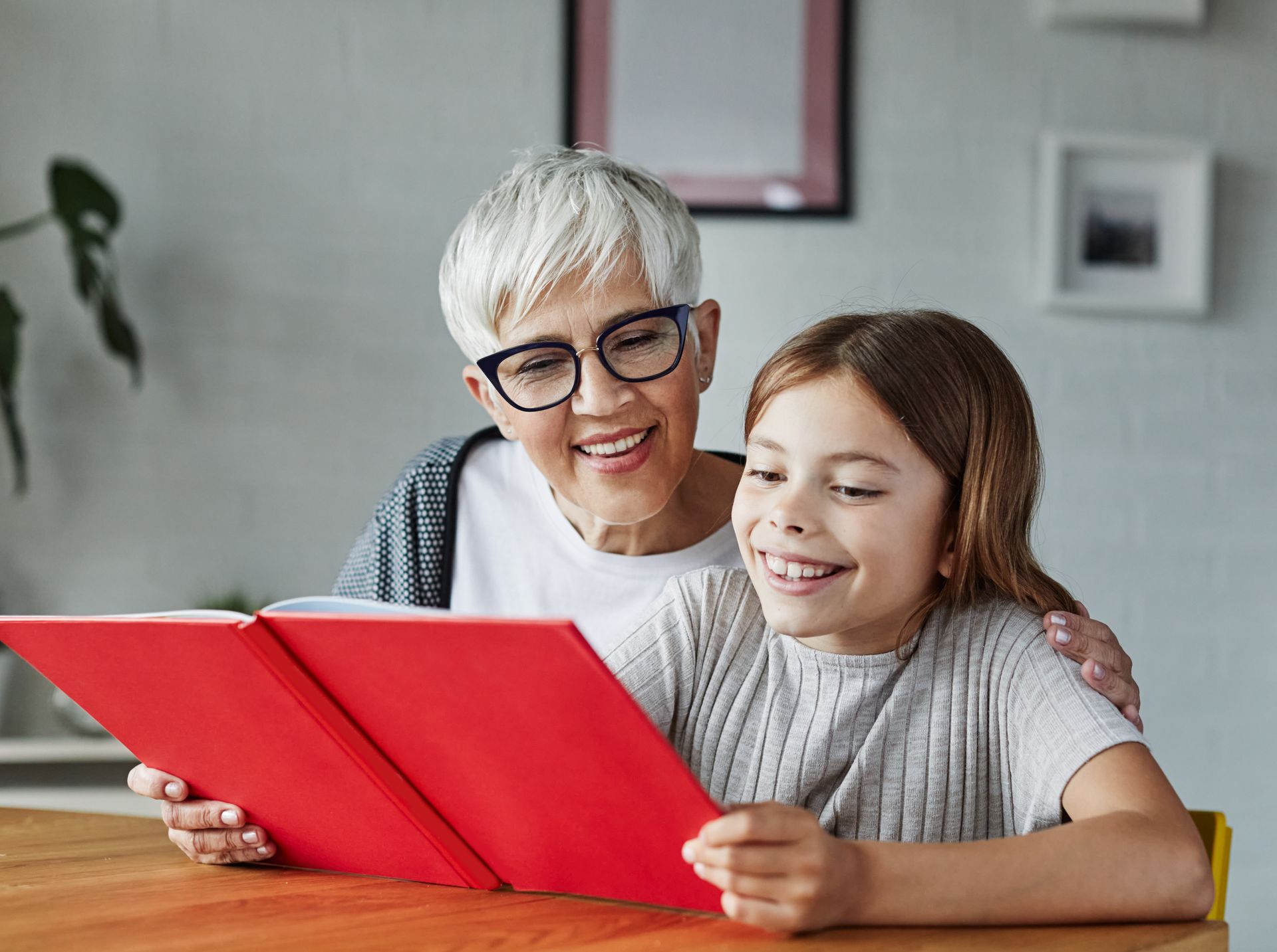 Grandmother wearing glasses and girl smiling while reading a red book at a table indoors.