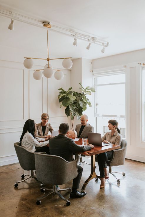 Businesspeople in a meeting around a round table, laptop visible. Brightly lit room with a large plant.