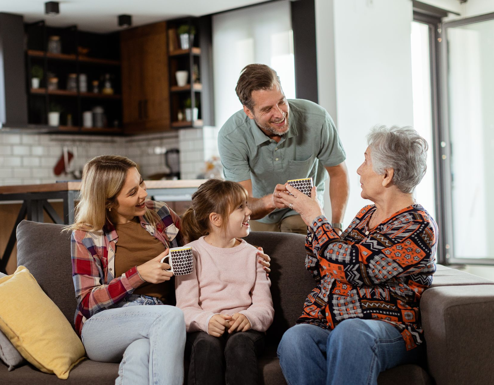 Family gathered on a couch, holding mugs, smiling at each other in a living room.