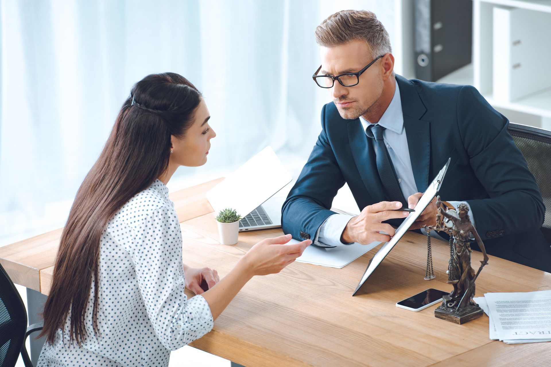 Woman speaking to a man in a suit, both looking at a document. They are at a desk with a law statue.