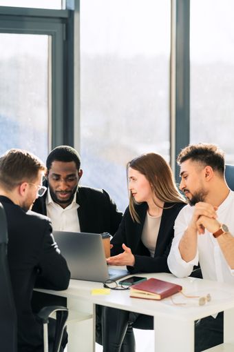 Four people in a meeting, looking at a laptop. They are in a modern office with large windows.